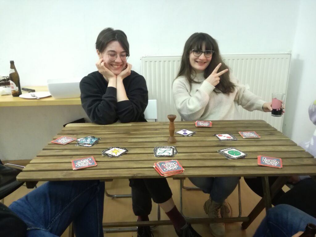 Auf dem Tisch liegen Spielutensilien vom Kartenspiel Jungle Speed, mehrere Menschen sitzen um den Tisch herum, in der Bildmitte zwei lächelnde Mitspielerinnen.