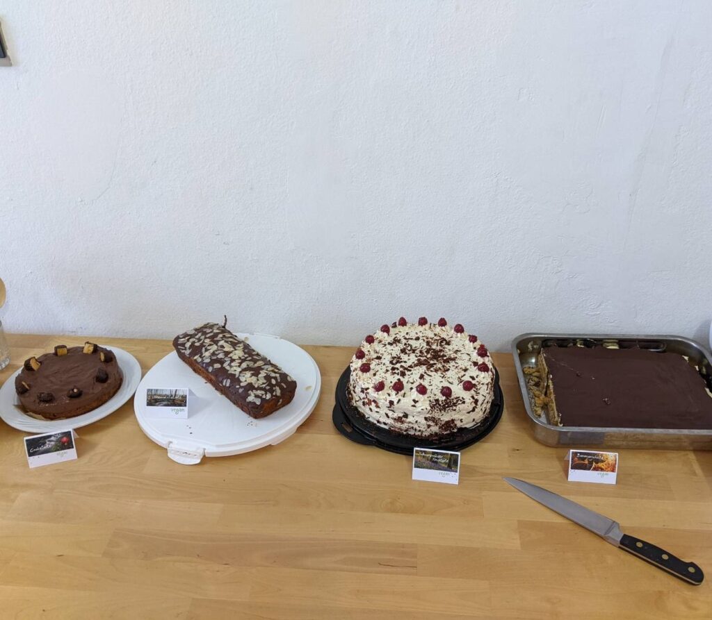 Auf einem Tisch sind nebeneinader vier vegane Kuchen samt Infokärtchen dazu präsentiert.